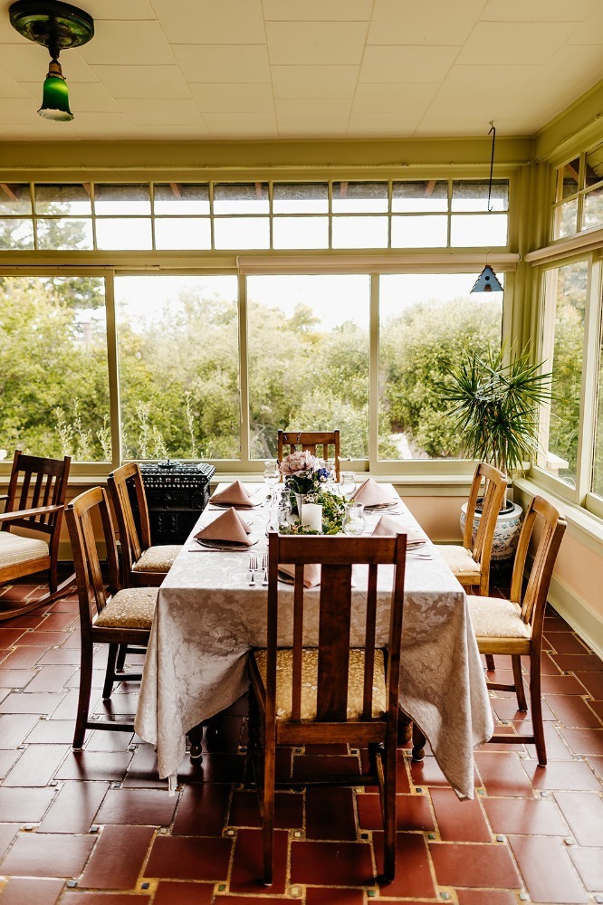 A dining area with a table set for a meal, surrounded by windows and plants.
