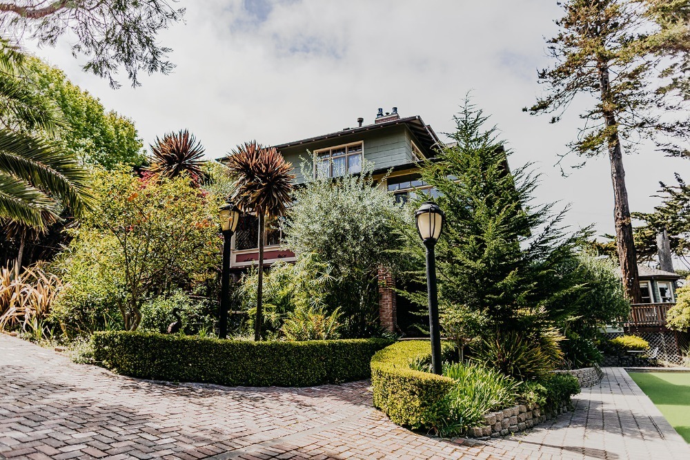 A large house with a green exterior, surrounded by lush gardens and trees, with a brick pathway leading to the entrance.