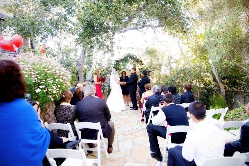 A wedding ceremony taking place outdoors with a floral arch, surrounded by trees and gardens.